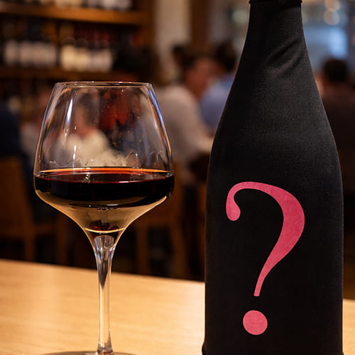 Verre de vin devant une bouteille masquée par une housse noire avec un point d’interrogation rose.
